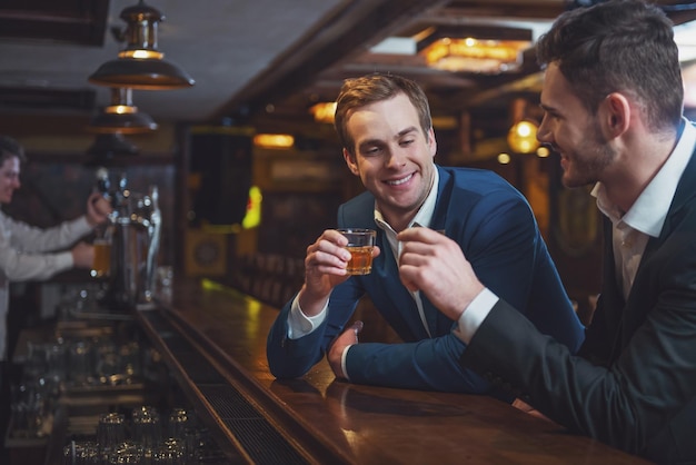 Zwei junge Geschäftsleute in Anzügen lächeln und klirren mit alkoholischen Getränken, während sie an der Bartheke im Pub sitzen