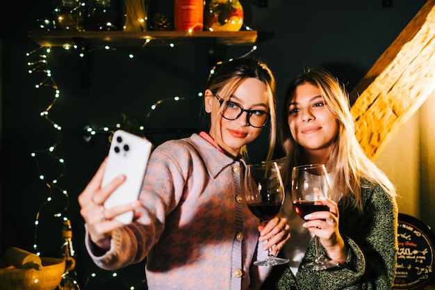 Zwei junge glückliche Frauen, die festliches Foto-Selfie nahe Weihnachtsbaum machen und Wein trinken.