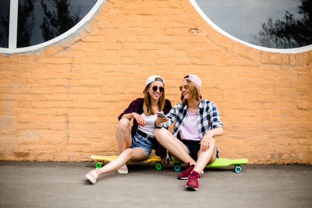 Zwei junge glückliche Freundinnen in Hipster-Outfit sitzen auf Longboards und machen Selfie am Telefon.