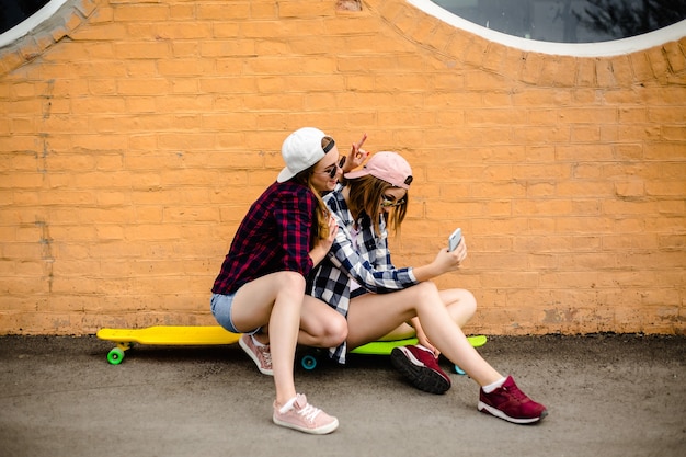 Zwei junge glückliche Freundinnen in Hipster-Outfit sitzen auf Longboards und machen Selfie am Telefon.