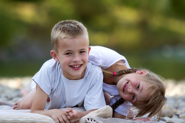 Zwei junge glückliche nette blonde lächelnde Kinder, Junge und Mädchen, Bruder und Schwester, die auf Kiesstrand auf unscharfer heller sonniger Sommertageszene umfasst werden. Freundschaft und perfektes Ferienkonzept.