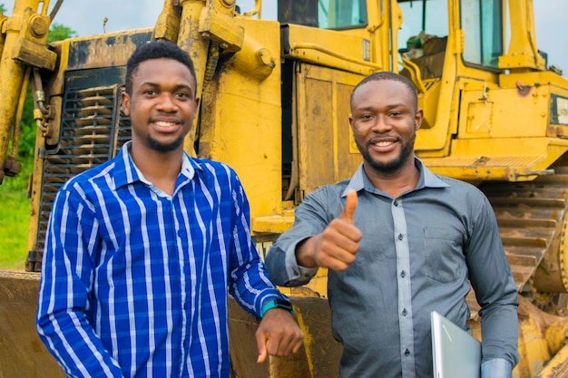 Zwei junge, gutaussehende afrikanische Bauunternehmer fühlen sich überwältigt, als sie neben einem Traktor stehen und Daumen hoch machen