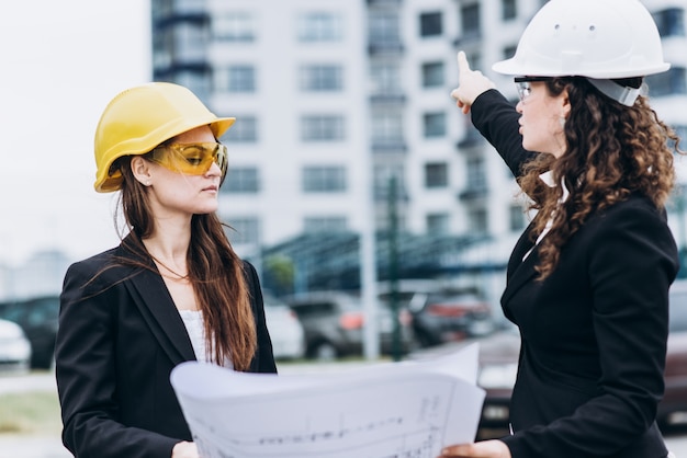 Zwei junge hübsche Business-Frauen Industrie-Ingenieure in Bau Helme auf einem Glas Gebäude Hintergrund. Bauplan, Architekt, Designer, erfolgreich