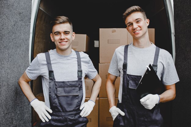 Zwei junge hübsche lächelnde Arbeiter in Uniform stehen vor dem Lieferwagen voller Kisten