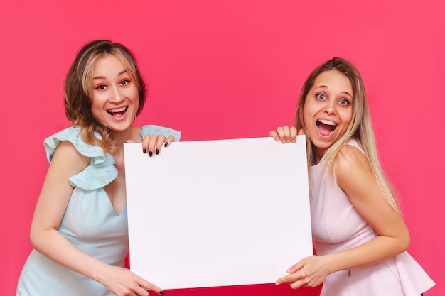 Zwei junge hübsche lächelnde, fröhliche Frauen-Promoter halten weiße Tafel an einer hellen rosa Wand