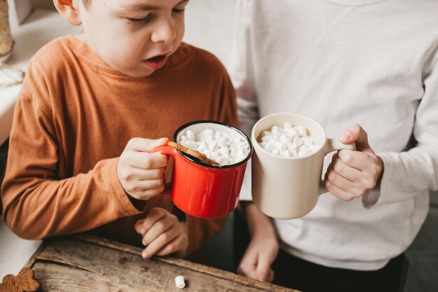 Zwei junge in Weihnachtskleidung, die Kakao mit Marshmallows in der Küche hält. Der Lebkuchenmann wird in Kakao gebadet. Weihnachtsstimmung. Weihnachtssüßigkeiten und Getränke, Textfreiraum