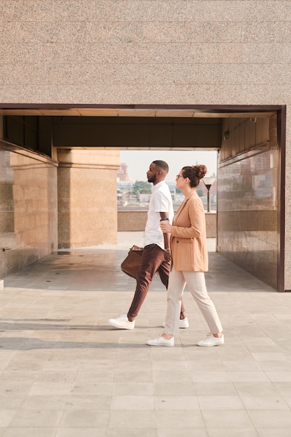 Zwei junge interkulturelle Makler in smarter Freizeitkleidung unterhalten sich, während sie die geflieste Straße entlang moderner Architektur im Freien entlang gehen