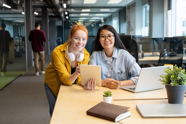 Zwei junge interkulturelle Managerinnen mit zahnigem Lächeln, die Sie ansehen, während sie zusammen über Präsentation durch Arbeitsplatz im Büro arbeiten