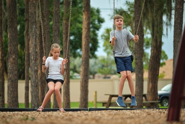 Zwei junge Kinder im Teenageralter, ein Mädchen und ein Junge, spielen zusammen auf Schaukeln im Freien an einem hellen, sonnigen Urlaubstag