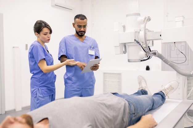 Zwei junge Kliniker in blauer Uniform diskutieren Online-Daten im Touchpad, während sie während der medizinischen Behandlung einem kranken Patienten zur Seite stehen