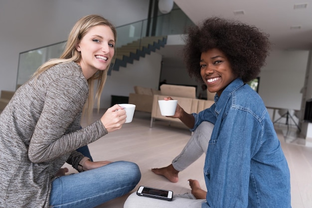 Zwei junge lachende multiethnische Frauen sitzen auf dem Boden und genießen beim Kaffeetrinken mit dem Smartphone im Luxushaus