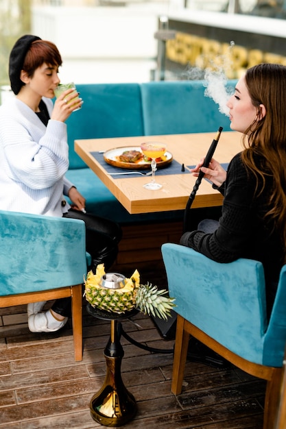 Zwei junge, lässig gekleidete Frauen trinken Cocktails und rauchen Wasserpfeife auf der Sommerterrasse in der Lounge