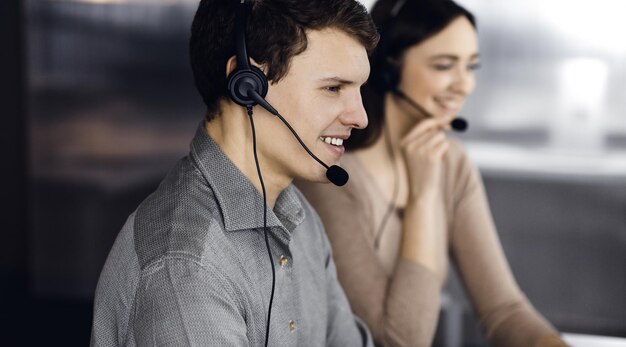 Zwei junge Leute in Kopfhörern sprechen mit den Kunden, während sie zusammen an dem Schreibtisch in einem modernen Büro sitzen, konzentrieren sich auf den Mann, Call-Center-Operatoren bei der Arbeit