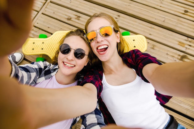 Zwei junge Mädchen in Hipster-Outfit machen Selfie beim Liegen mit auf Holzsteg.