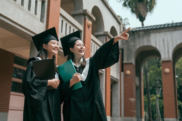 zwei junge mädchen studenten am abschlusstag, die ein lehrbuchstudium im ausland halten, beenden die universität. Frau zeigt mit dem Finger einen Freund lächelnd, lachend, plaudernd, in einem traditionellen roten Backsteinhaus.