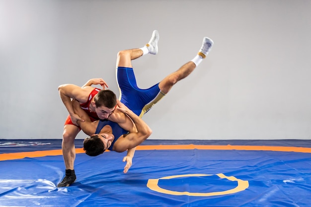 Zwei junge Männer in blauen und roten Wrestling-Strumpfhosen ringen und machen ein Suplex-Wrestling auf einem gelben Wrestling-Teppich in der Turnhalle