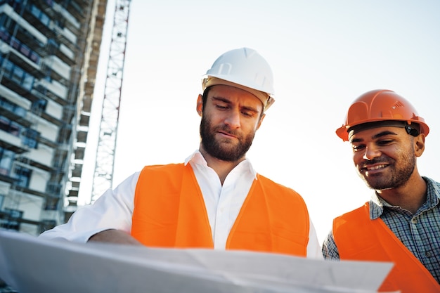 Zwei junge männliche Ingenieure in Uniform und Schutzhelmen, die auf der Baustelle arbeiten