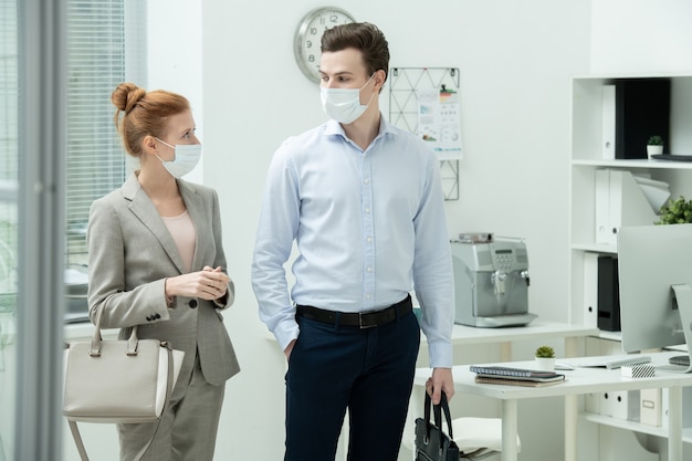 Zwei junge Manager in Schutzmasken und Abendgarderobe diskutieren After-Work-Pläne beim Verlassen des Büros und verbringen Zeit miteinander