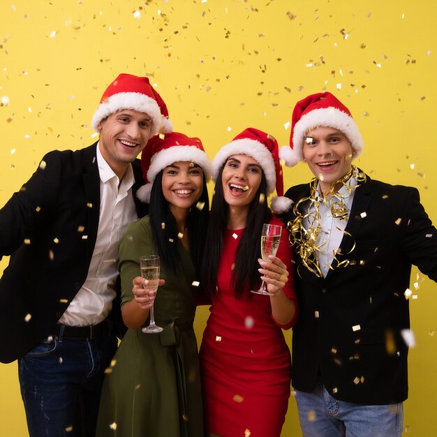 Zwei junge Paare in Weihnachtsmützen feiern eine Silvesterparty, lachen vor Freude, jubeln mit Sektgläsern und werfen buntes Konfetti