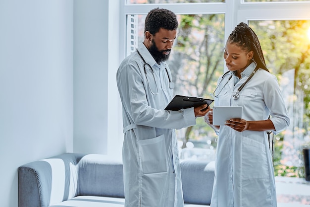 Zwei junge Ärzte in weißen Kitteln besprechen die Behandlung in der Krankenhauslobby und schauen auf das Tablet