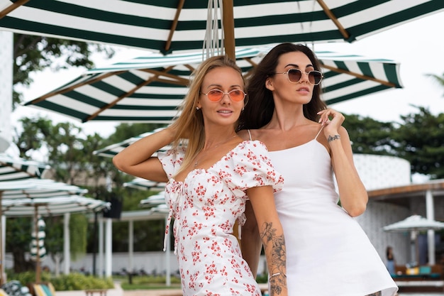 Zwei junge schöne Frauen in trendigem, elegantem Sommerkleid und Sonnenbrille posieren im Urlaub stilvoll und modisch auf Strandclub-Hintergrund
