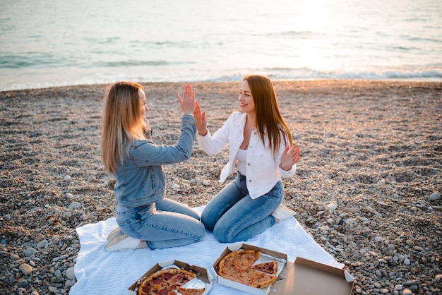 Zwei junge schöne Freundinnen, die Spaß mit Pizza am Strand über dem Meer im Freien im Sonnenlicht haben Sommerferienzeit Freundschaft Glück Schwestern verbringen Zeit zusammen an der Küste