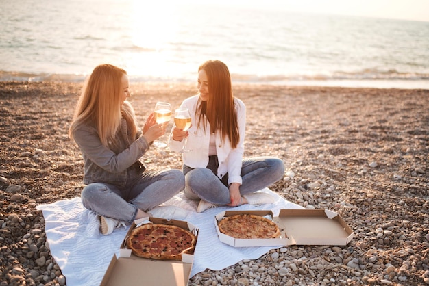 Zwei junge schöne Freundinnen, die Spaß mit Pizza und Wein am Strand über dem Meeresufer im Freien im Sonnenlicht haben. Sommerferienzeit. Freundschaft. Glück. Schwestern verbringen Zeit zusammen an der Küste.