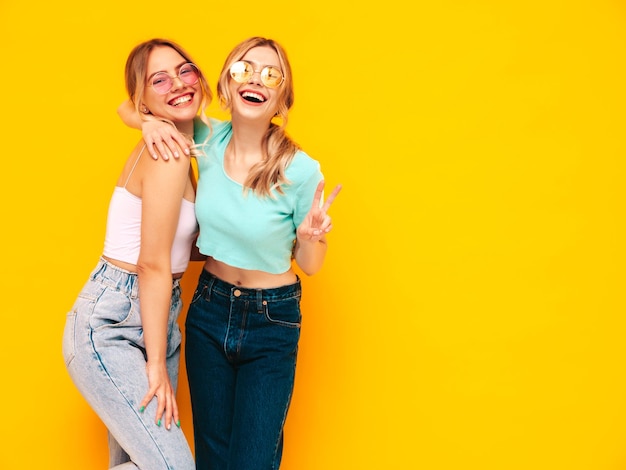 Zwei junge schöne lächelnde blonde hipster-frauen in trendiger sommerkleidung sexy sorglose frauen posieren in der nähe der gelben wand im studio positive models, die spaß haben fröhlich und glücklich in sonnenbrillen