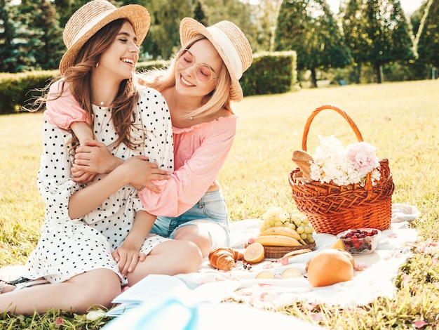 Zwei junge schöne lächelnde Frauen in trendigem Sommerkleid und Hüten. Sorglose Frauen, die draußen Picknick machen.