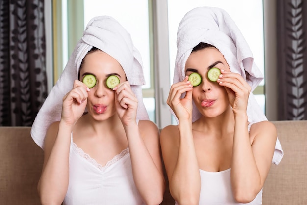 Zwei junge schöne lächelnde Freundinnen mit Handtüchern auf dem Kopf tragen Gurkenringe auf ihre Augen auf und machen eine kosmetische Maske