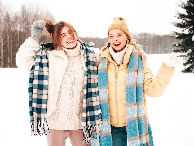 Zwei junge schöne lächelnde Hipster-Frau in trendiger warmer Kleidung und Schals. Sorglose Frauen, die auf der Straße im Park posieren. Positive pure Models, die Spaß im Schnee haben. Wintermomente genießen. Weihnachtskonzept