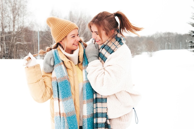 Zwei junge schöne lächelnde Hipster-Frau in trendiger warmer Kleidung und Schals. Sorglose Frauen, die auf der Straße im Park posieren. Positive pure Models, die Spaß im Schnee haben. Wintermomente genießen. Weihnachtskonzept