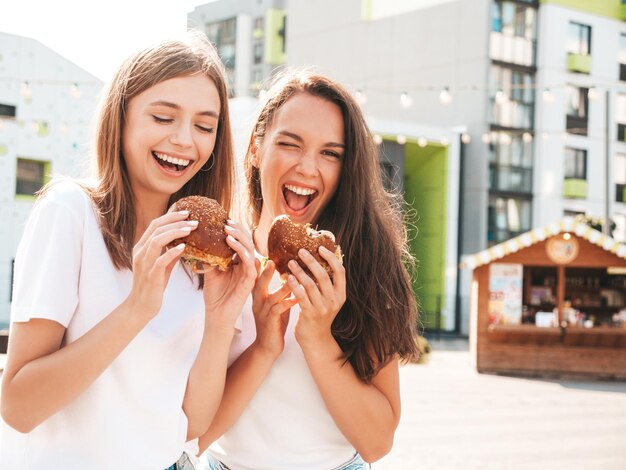 Zwei junge schöne lächelnde Hipster-Frauen im trendigen Sommer gleiche Kleidung Sexy sorglose Frauen posieren auf der StraßePositive Models, die Spaß mit Sonnenbrillen habenHolding saftiger Burger und Hamburger essen