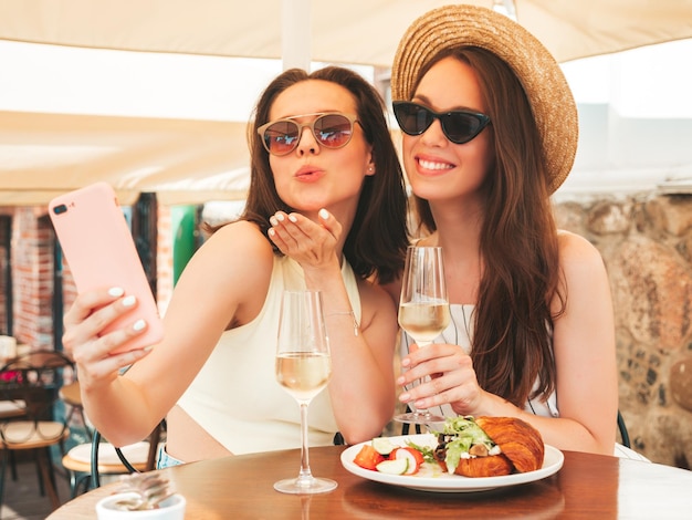 Zwei junge schöne lächelnde Hipster-Frauen in trendiger SommerkleidungSorglose Frauen posieren im Veranda-Café auf der StraßePositive Models trinken Weißwein im HutSelfie-Fotos machen