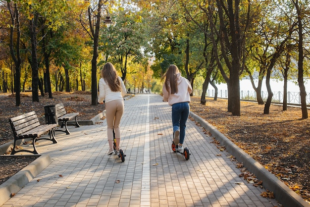 Zwei junge schöne Mädchen fahren an einem warmen Herbsttag Elektroroller im Park.