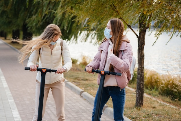 Zwei junge schöne Mädchen in Masken fahren an einem warmen Herbsttag Elektroroller im Park. Im Park spazieren gehen.