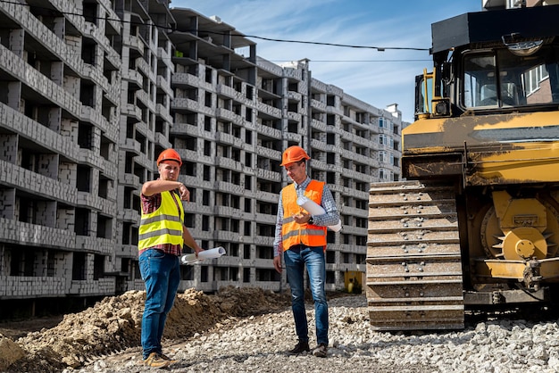 Zwei junge starke Arbeiter in Uniform und Helm stehen neben einem Grader auf einer Baustelle. Baukonzept