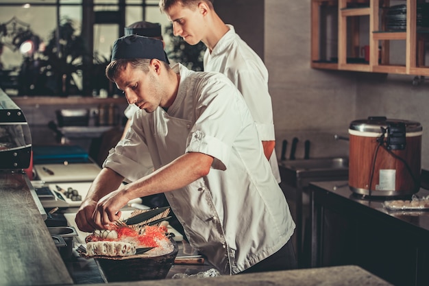 Zwei junge weiße Köche in weißer Uniform schmücken fertiges Gericht im Restaurant