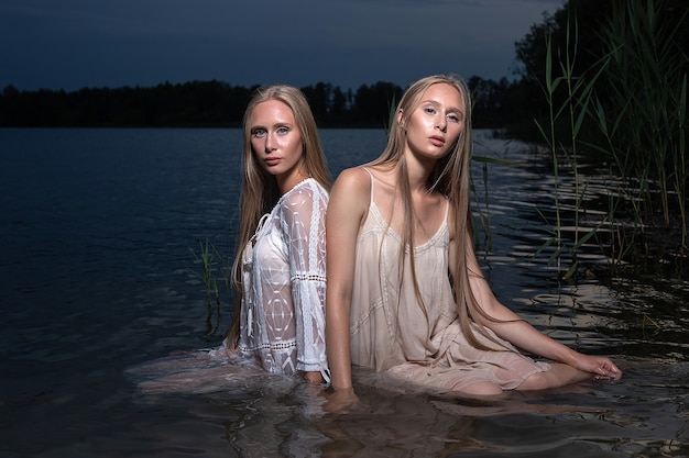 Zwei junge Zwillingsschwestern mit langen blonden Haaren, die in der Sommernacht in hellen Kleidern im Wasser des Sees aufwerfen. im Freien abends Fotosession