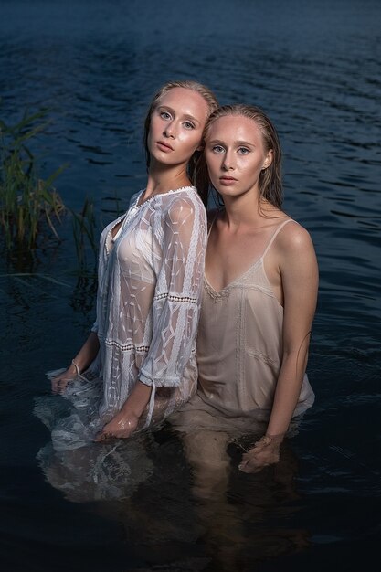 Zwei junge Zwillingsschwestern mit langen blonden Haaren, die in hellen Kleidern im Wasser des Sees in der Sommernacht aufwerfen