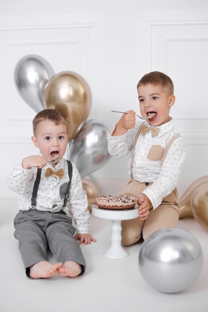 Zwei Jungen feiern Geburtstag, Kinder haben eine B-Day-Party. Geburtstagstorte mit Kerzen und Luftballons. Fröhliche Kinder essen Kuchen, Feiern, weißes minimalistisches Interieur.