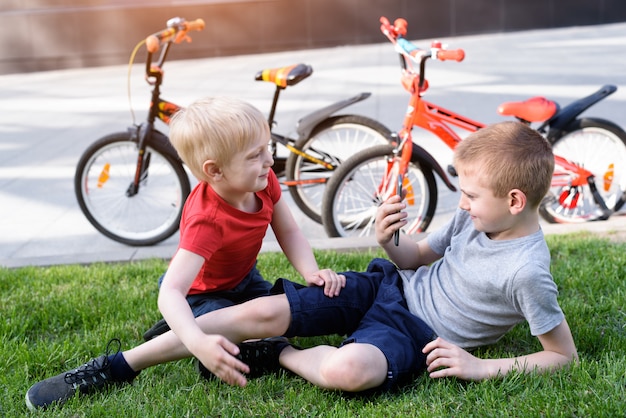 Zwei Jungen werden auf einem Smartphone fotografiert, während sie auf dem Gras sitzen. Erholung nach dem Radfahren, Fahrräder im Hintergrund