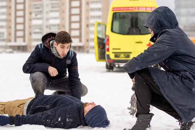 Zwei Kerle sitzen auf Kniebeugen durch kranken oder bewusstlosen jungen Mann, der im Schnee liegt