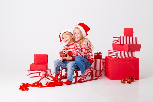 Zwei Kinder, ein Junge und ein Mädchen in Pullovern und Hüten Santa, sitzen auf einem Schlitten, um Geschenke zu tragen. Studio, weißer Hintergrund, Platz für Text