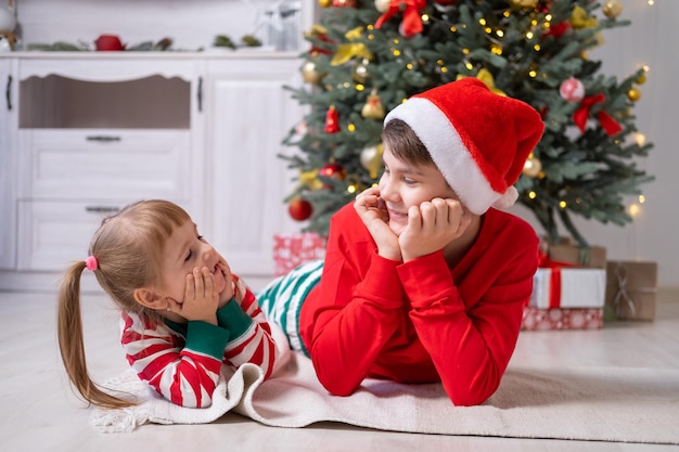 Zwei Kinder in Weihnachtspyjamas mit Geschenkboxen, die zu Hause unter dem Weihnachtsbaum liegen