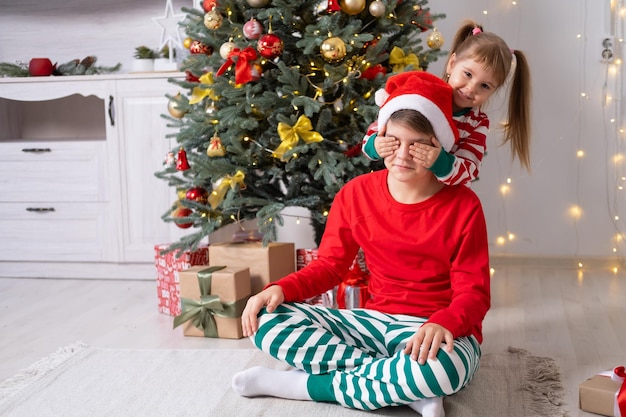 Zwei Kinder in Weihnachtspyjamas mit Geschenkboxen, die zu Hause unter dem Weihnachtsbaum liegen