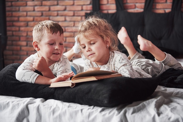 Zwei Kinder liegen auf einem großen Bett und lesen ein interessantes Buch. Sie tragen den gleichen Pyjama