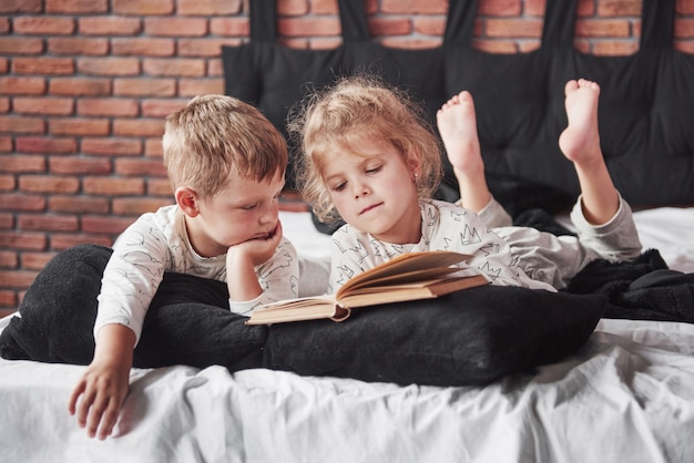 Zwei Kinder liegen auf einem großen Bett und lesen ein interessantes Buch. Sie tragen den gleichen Pyjama