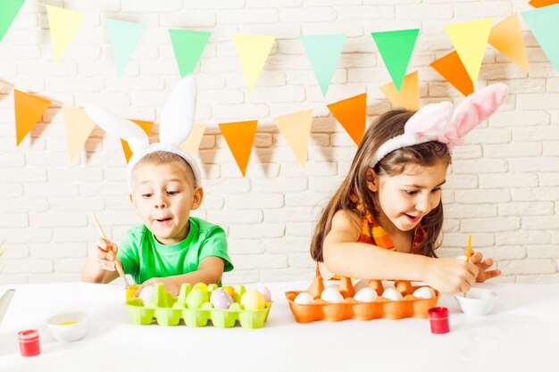 Zwei Kinder mit Osterhasenohren, die zu Hause bunte Ostereier malen. Der kleine Junge und das Mädchen bereiten sich auf Ostern vor