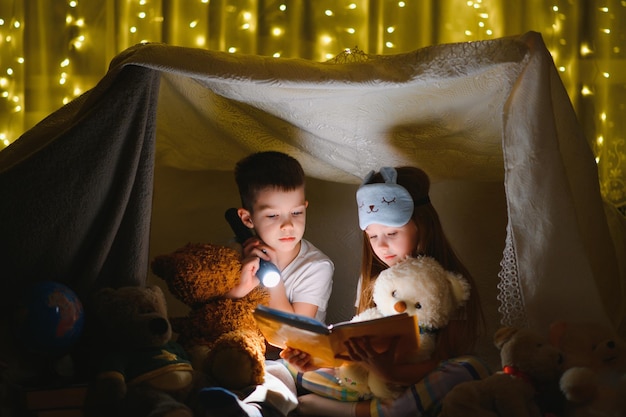 Zwei Kinder mit Taschenlampe lesen ein Buch unter einer Decke als Zelt